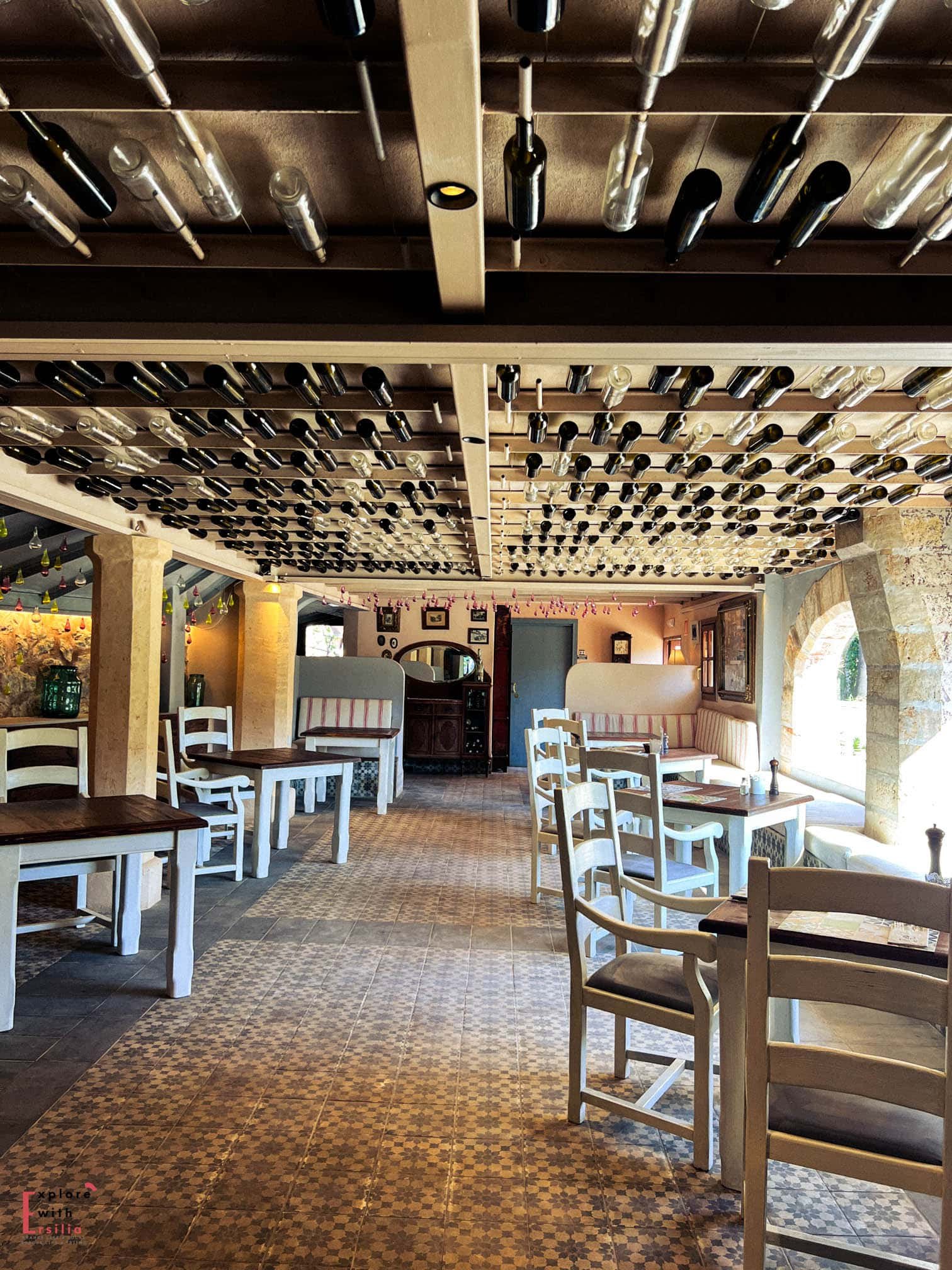 Restaurant interior at Finca Can N'ai featuring a dramatic ceiling installation of wine bottles, white-painted rustic furniture, and traditional patterned tile floors. Stone arches and country-style benches with striped cushions complete the Mediterranean farmhouse atmosphere.