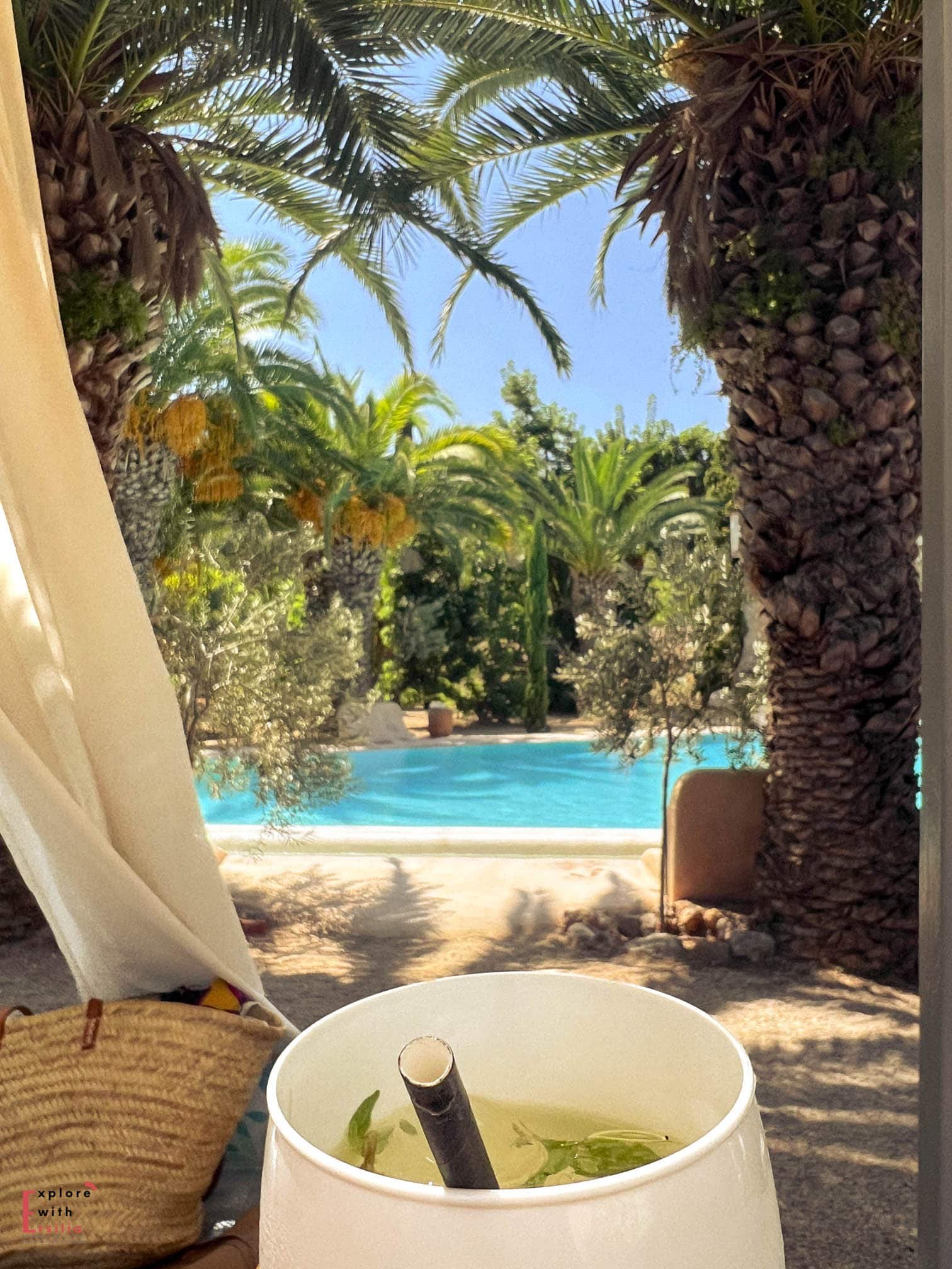 A glass of Hugo/St Germain cocktail with fresh mint leaves in a white wine glass, viewed from a cabana with the turquoise pool and palm trees of Finca Can N'ai in the background. A straw beach bag sits nearby.