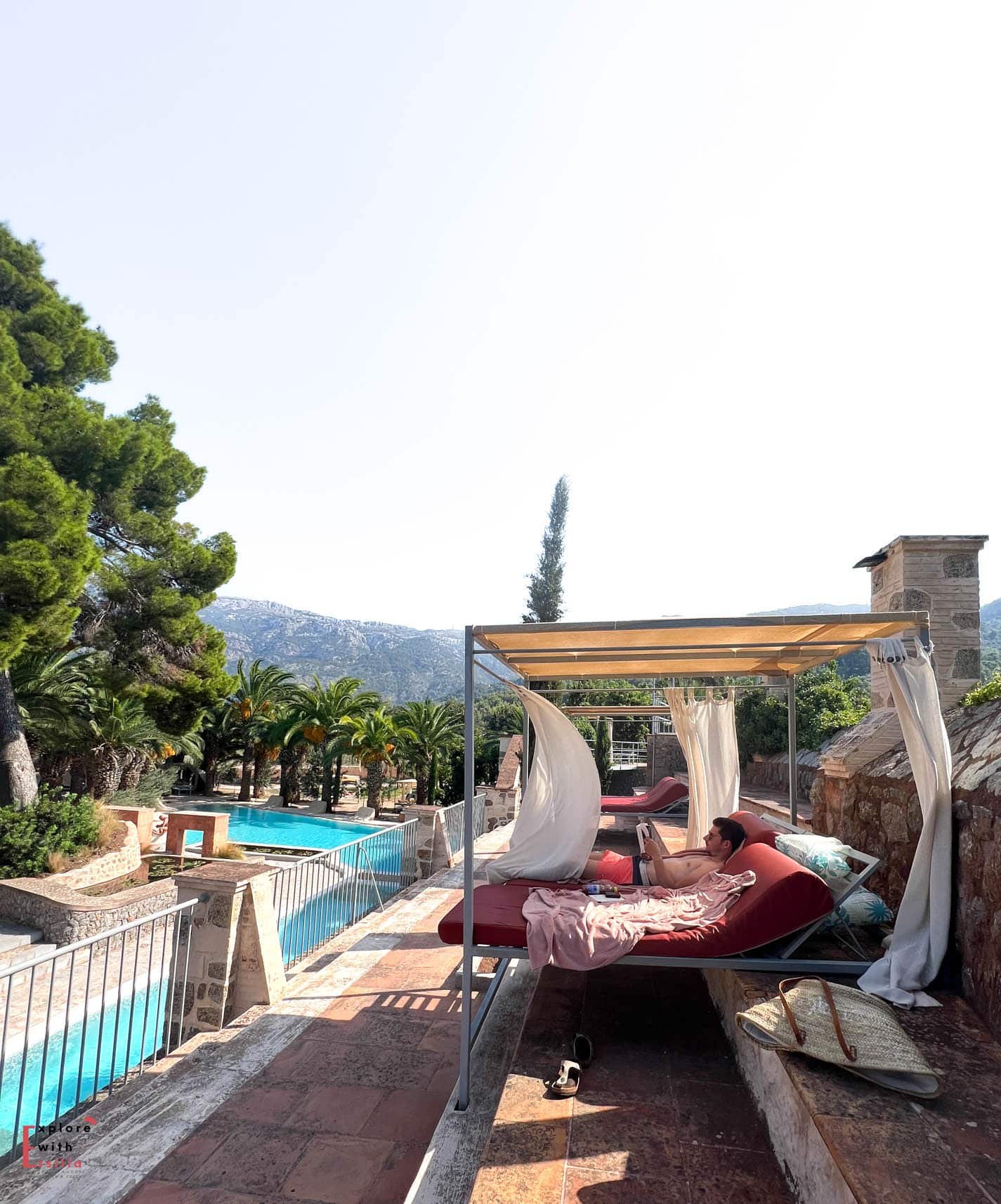 Poolside relaxation area at Finca Can N'ai featuring a covered daybed with flowing white curtains overlooking the multi-level swimming pool, palm trees, and Serra de Tramuntana mountains in the background