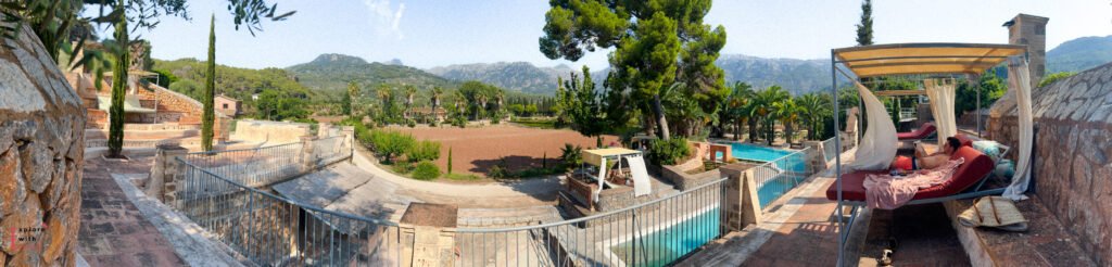 Panoramic view of Finca Can N'ai's grounds showing the swimming pool, terraced gardens, and stone buildings against the backdrop of Serra de Tramuntana mountains, with a covered daybed lounging area in the foreground.