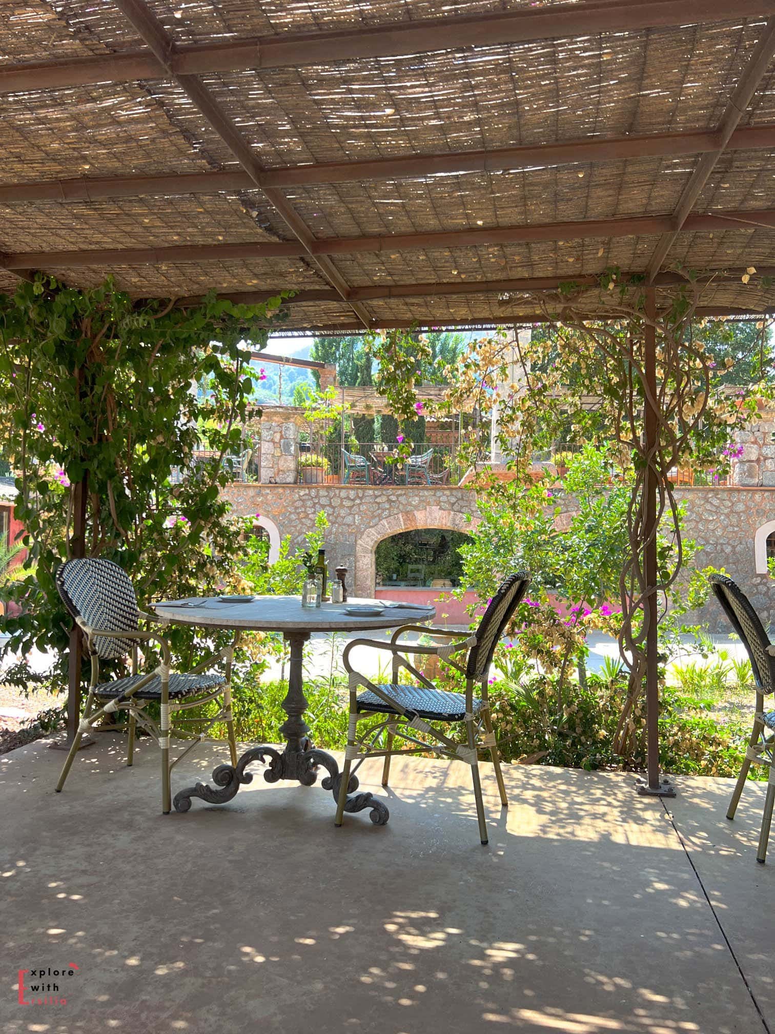 Outdoor dining area at Finca Can n'ai in Mallorca, sheltered by a traditional reed pergola casting dappled shadows. A bistro table with ornate cast iron base and woven chairs sits among climbing vines and bougainvillea, with a stone arch terrace visible in the background.