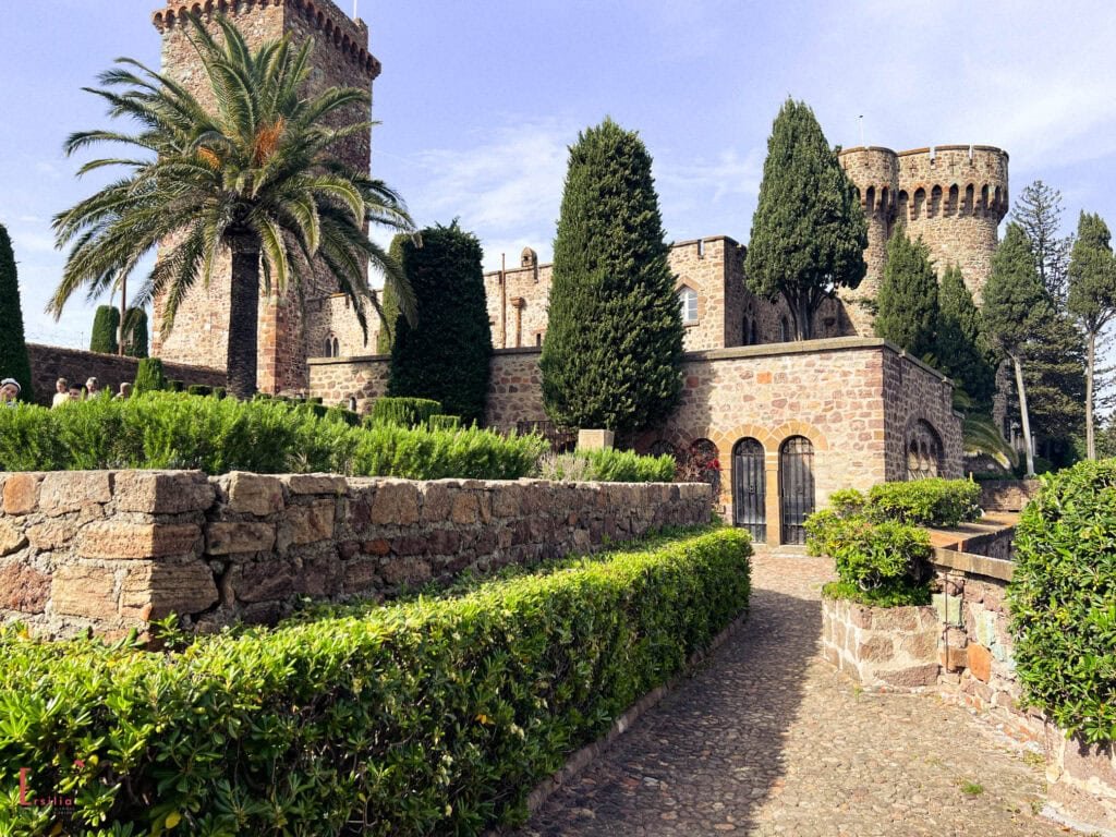 Medieval Château de la Napoule with stone towers, lush Mediterranean gardens featuring palm trees, cypress trees, and manicured hedges, connected by a cobblestone pathway along the ancient stone walls under clear blue skies on the French Riviera.