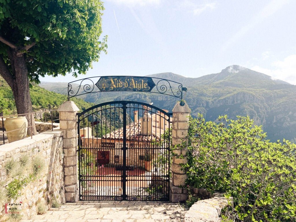 Wrought iron gate with 'Le Nid d'Aigle' (Eagle's Nest) sign at the entrance of a viewpoint in Gourdon village, with dramatic mountain views of the French Alps in the background and Mediterranean vegetation framing the scene.