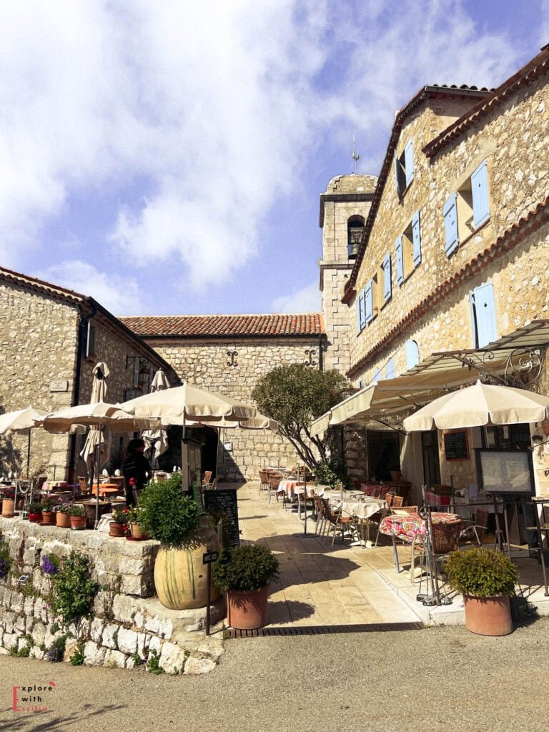 A charming outdoor terrace of La Taverne Provençale restaurant in Gourdon, featuring cream-colored umbrellas, stone walls, and potted plants. The medieval church bell tower rises above the honey-colored stone buildings with pale blue shutters, all under a bright blue sky with wispy clouds