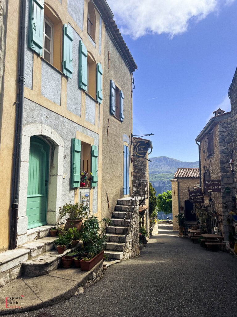 Street scene in Gourdon village featuring a stone house with mint green shutters and door, stone steps leading up to shops, and potted plants decorating the entrance. The narrow street offers stunning views of the mountains in the background.