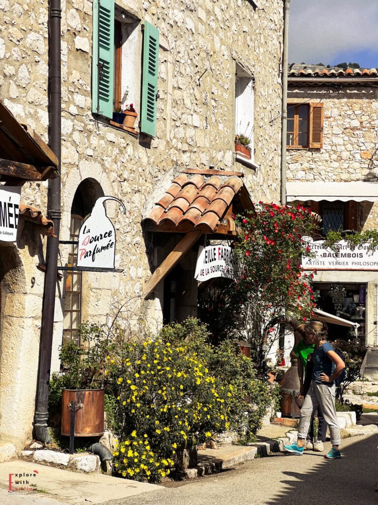 Charming storefront in Gourdon showing 'La Source Parfumée' perfume shop with stone walls, mint green shutters, and terracotta roof tiles. Yellow wildflowers and red climbing roses decorate the entrance, with vintage-style shop signs and artisan displays.