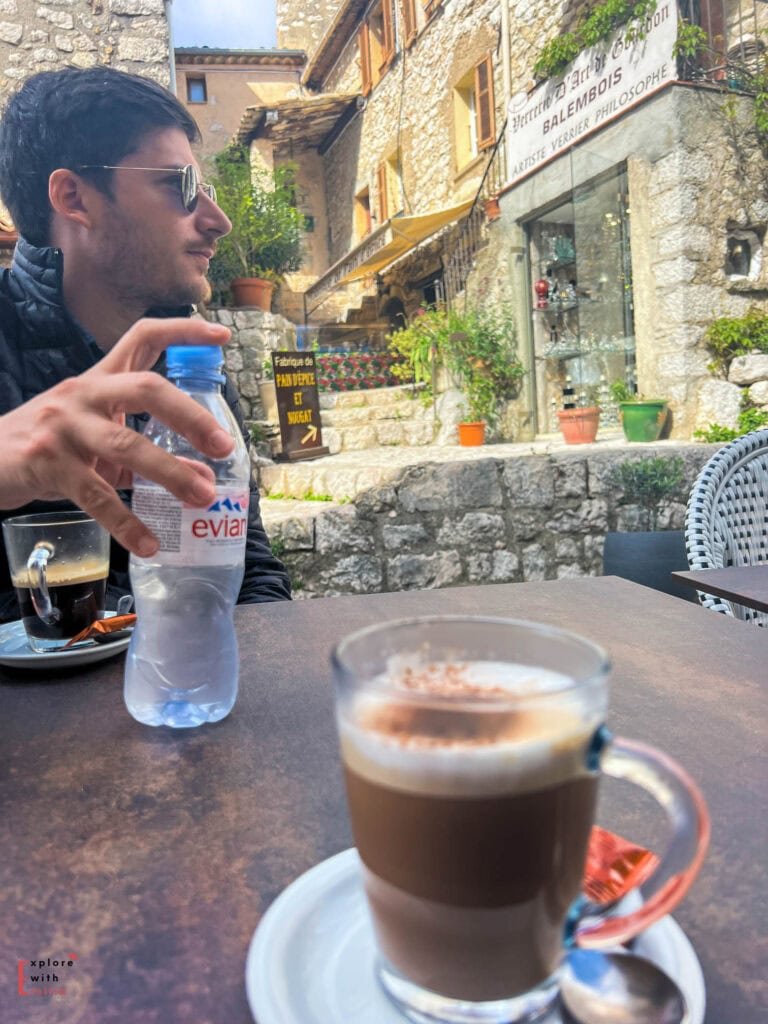 Layered coffee drink at an outdoor café in Gourdon village, with historic stone buildings and artisan shops visible in the background. A bottle of Evian water and a person in sunglasses sit at the café table overlooking the medieval village scene.