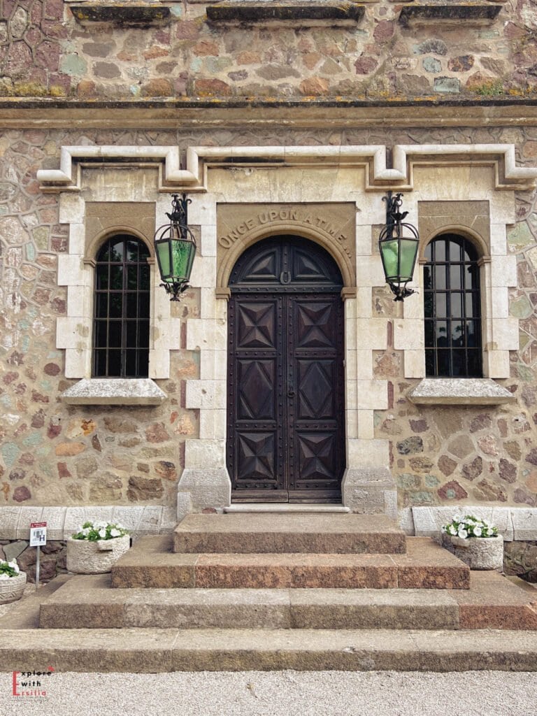 Grand entrance at Château de la Napoule featuring a dark wooden door with geometric panels, inscribed with 'Once Upon a Time' above. Two green glass lanterns flank the doorway, with stone windows on either side and white flower planters decorating the steps.