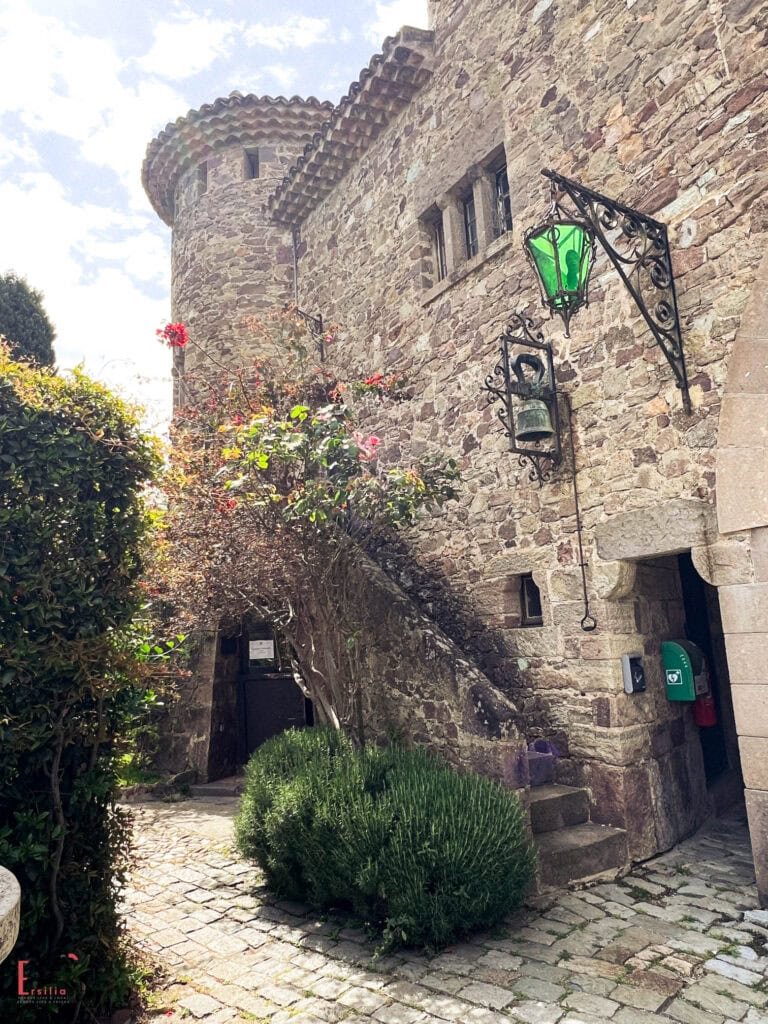 Medieval entrance to Château de la Napoule featuring a round stone tower with terracotta roof tiles, decorative iron lantern with green glass, stone stairway, flowering bougainvillea vine, and Mediterranean herbs growing along the cobblestone pathway.