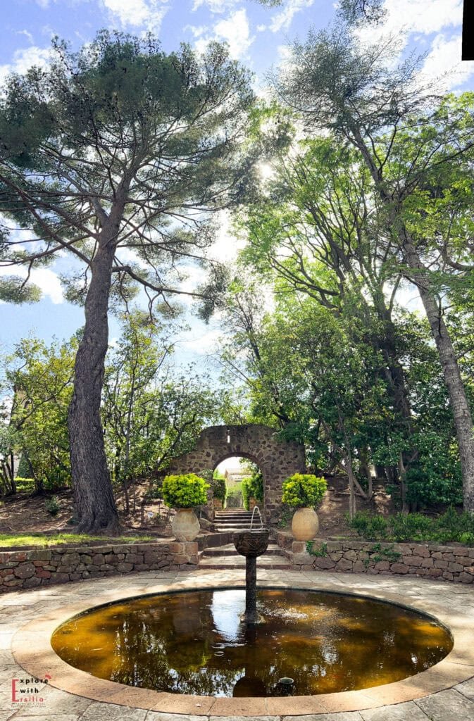 Garden fountain in Château de la Napoule's grounds featuring a circular stone basin with central fountain, framed by a stone archway in the background. Tall pine trees provide shade while large terracotta planters with boxwood flank the archway steps.