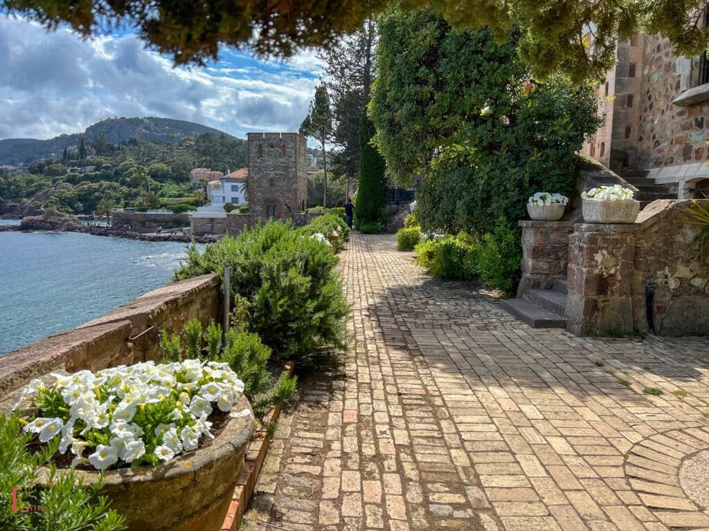 Coastal stone pathway along Château de la Napoule gardens featuring white flower planters, lush Mediterranean vegetation, historic stone architecture, and panoramic views of the azure sea, distant hills, and the castle's medieval watchtower.