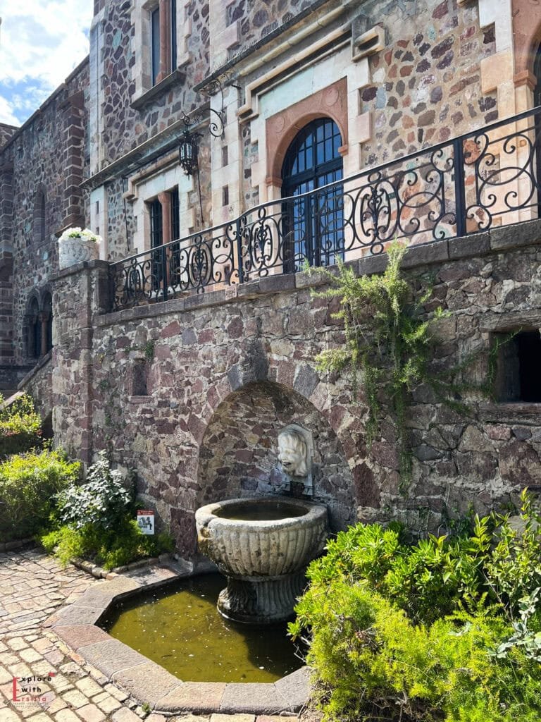 Historic stone fountain with a face sculpture at Château de la Napoule featuring multicolored stone walls, wrought iron balconies, arched windows, and lush Mediterranean vegetation growing along the medieval castle exterior.