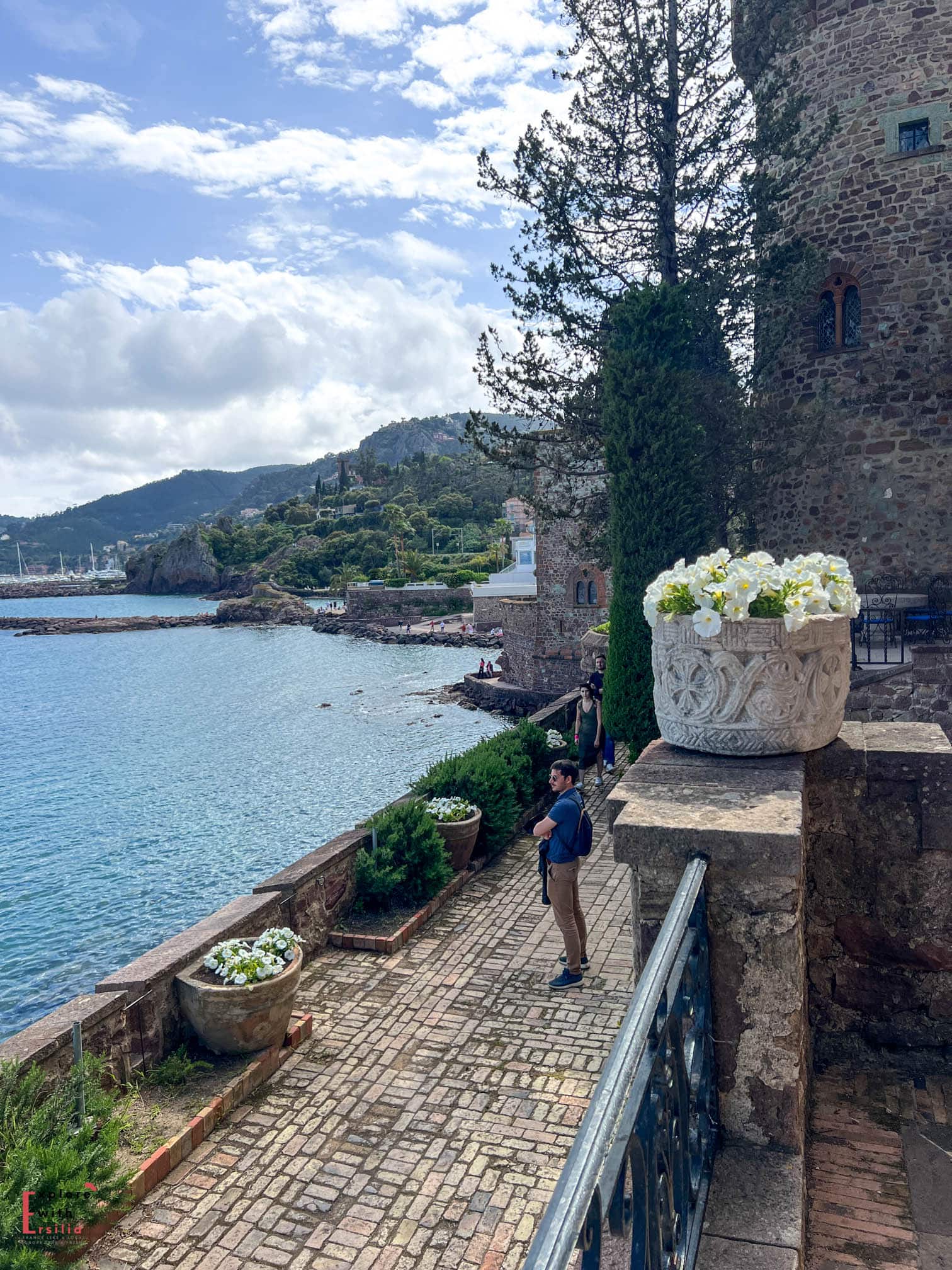 Seaside walkway at Château de la Napoule on the French Riviera, featuring a stone-paved path with ornate white planters filled with flowers. A medieval stone castle wall runs alongside the path, with the Mediterranean Sea and coastal mountains creating a dramatic backdrop.