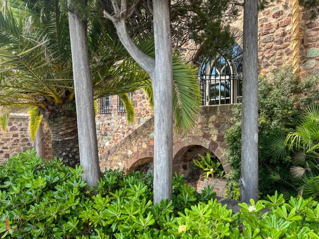 Mediterranean garden at Château de la Napoule with palm fronds, silver-trunked trees, lush green vegetation, and a stone archway framing an ornate wrought iron window overlooking the multicolored stone walls of the medieval castle.