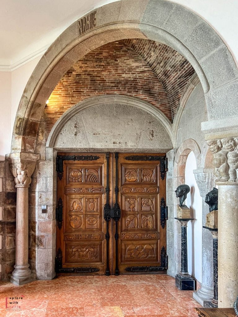 Intricately carved wooden doors with animal motifs inside a stone archway at Château de la Napoule, featuring the name 'MARIE' carved into the keystone, decorated with ornate hinges, flanked by marble columns and antique busts, showcasing the castle's unique blend of Gothic, Romanesque, and Saracen architectural styles.