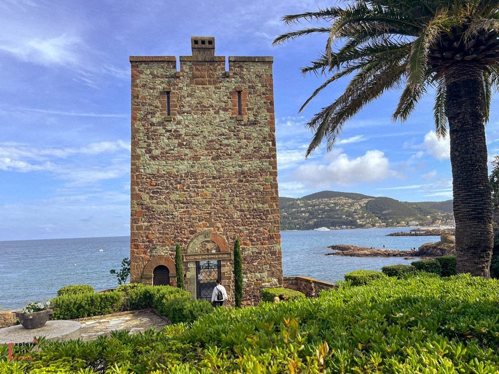 Medieval watchtower of Château de la Napoule overlooking the Mediterranean Sea, featuring stone masonry, crenellated top, arched entrance with decorative ironwork gate, surrounded by lush coastal gardens and palm trees on the French Riviera.