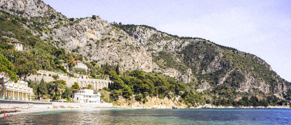 Panoramic view of Eze beach, showing a small sandy cove nestled beneath dramatic limestone cliffs, with Belle Époque villas and a historic stone viaduct visible along the mountainside, and the Mediterranean's turquoise waters in the foreground