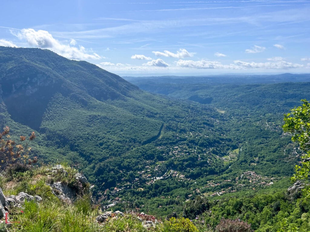 Panoramic view from Gourdon village overlooking a deep valley in the French Pre-Alps, with forested mountainsides, small villages nestled in the greenery, and distant hills fading into the horizon under a bright blue summer sky.