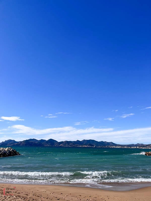 A vibrant seascape of the Lérins Islands off Cannes, featuring turquoise Mediterranean waters, rolling waves meeting a sandy beach, and a rugged mountain backdrop under a brilliant blue sky with wispy clouds.