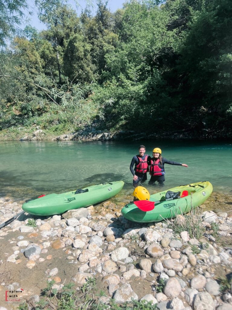 Two kayakers wearing red life vests and safety gear stand in turquoise waters next to their green kayaks resting on a rocky riverbank. The scene is framed by lush vegetation and trees along the riverbank under clear skies.