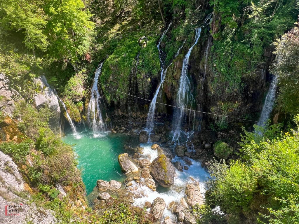 Multiple cascading waterfalls pour down moss-covered cliffs into a stunning turquoise pool at Les Gorges du Loup. The falls are surrounded by lush green vegetation, with large boulders scattered in the emerald waters below. A thin cable or wire stretches across the gorge above the falls.