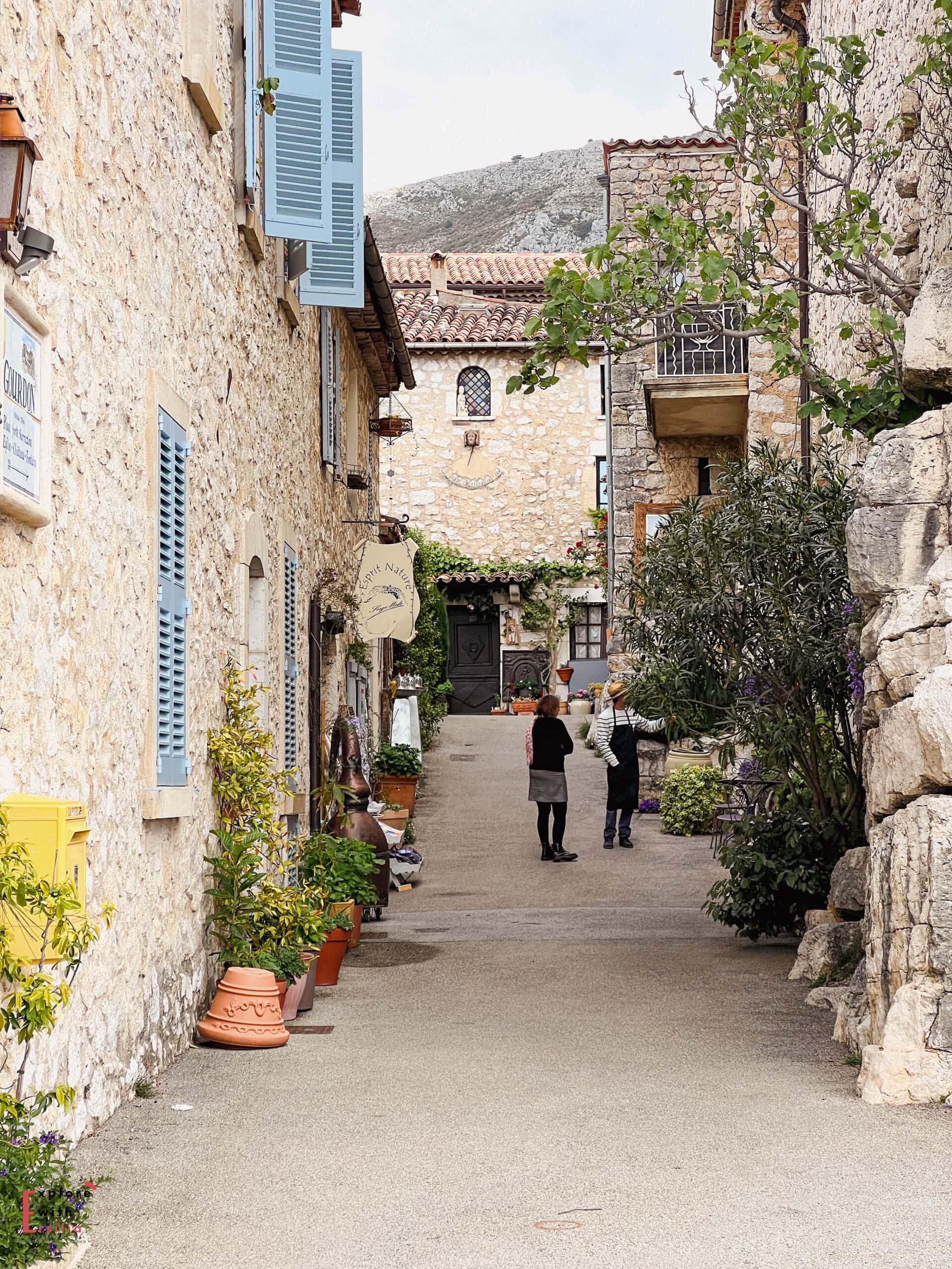 A charming cobblestone street in medieval Gourdon village, lined with honey-colored stone buildings featuring blue shutters and terracotta planters. Two people stand chatting along the narrow lane decorated with potted plants and climbing vines, while mountain views peek through in the background