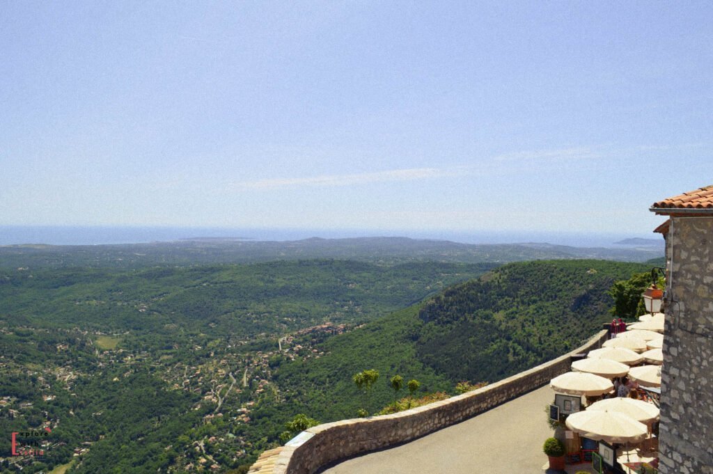 Alt text: "Panoramic view from La Taverne Provençale's terrace in Gourdon, showing a dramatic vista of the French Riviera countryside. The stone terrace with cream umbrellas overlooks a vast green valley dotted with villages, stretching all the way to the Mediterranean Sea on the horizon under a clear blue sky.