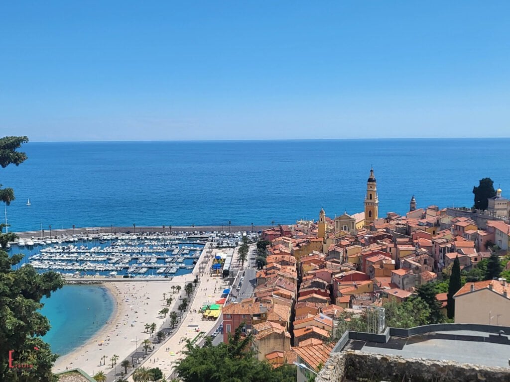 Panoramic view of Menton on the French Riviera, showing the colorful Old Town with orange terracotta rooftops, the prominent bell tower of Basilica St. Michel, a sandy beach along the promenade, marina filled with white boats, and the deep blue Mediterranean Sea stretching to the horizon under clear azure skies.