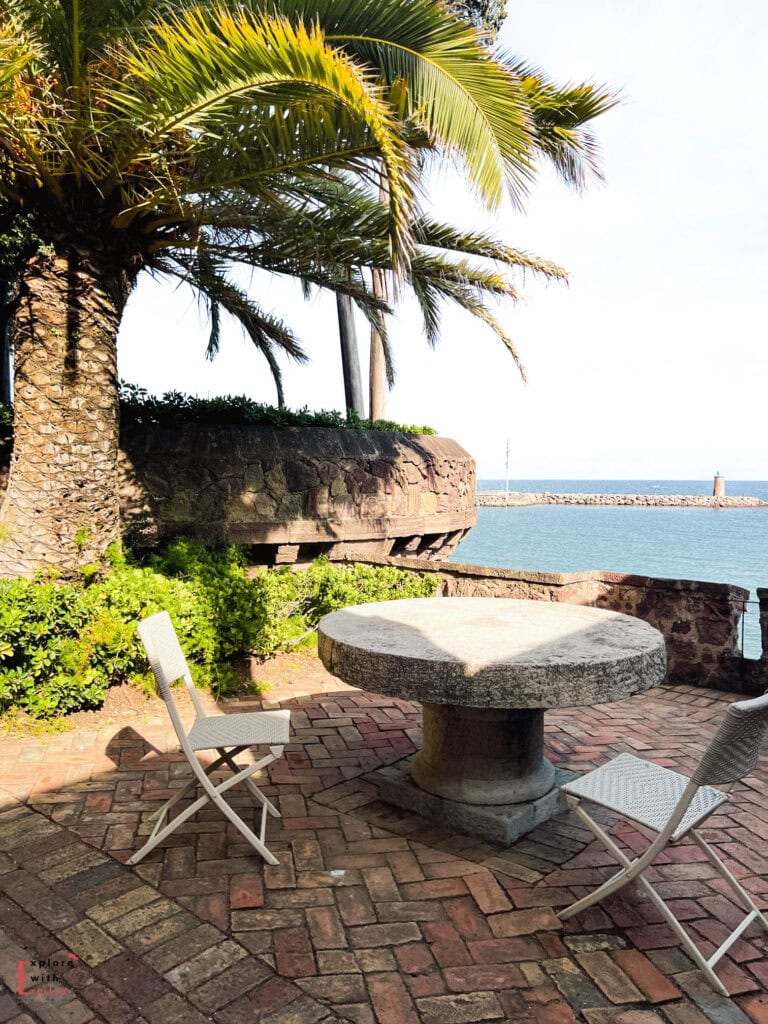 Seaside terrace with stone table and chairs overlooking the Mediterranean Sea, shaded by palm trees, with a historic stone wall and small lighthouse visible in the distance along the French Riviera.