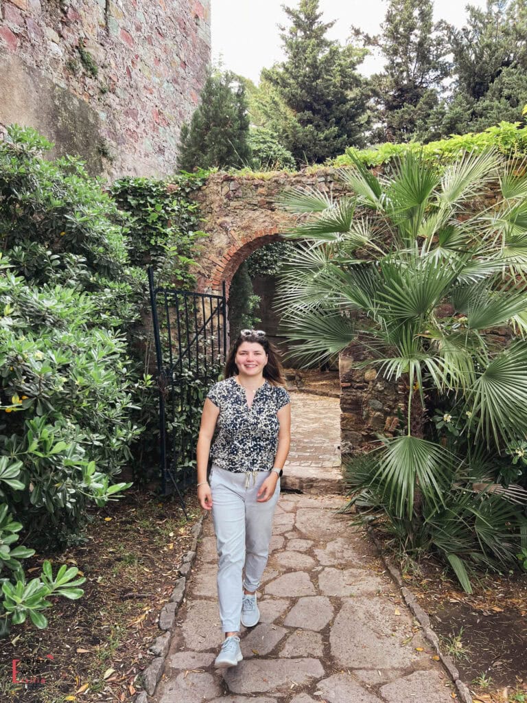 Garden pathway at Château de la Napoule featuring an ivy-covered stone archway, cobblestone walkway, fan palms, cypress trees, and Mediterranean plants, with a visitor walking through this lush passageway surrounded by the castle's historic stone walls.