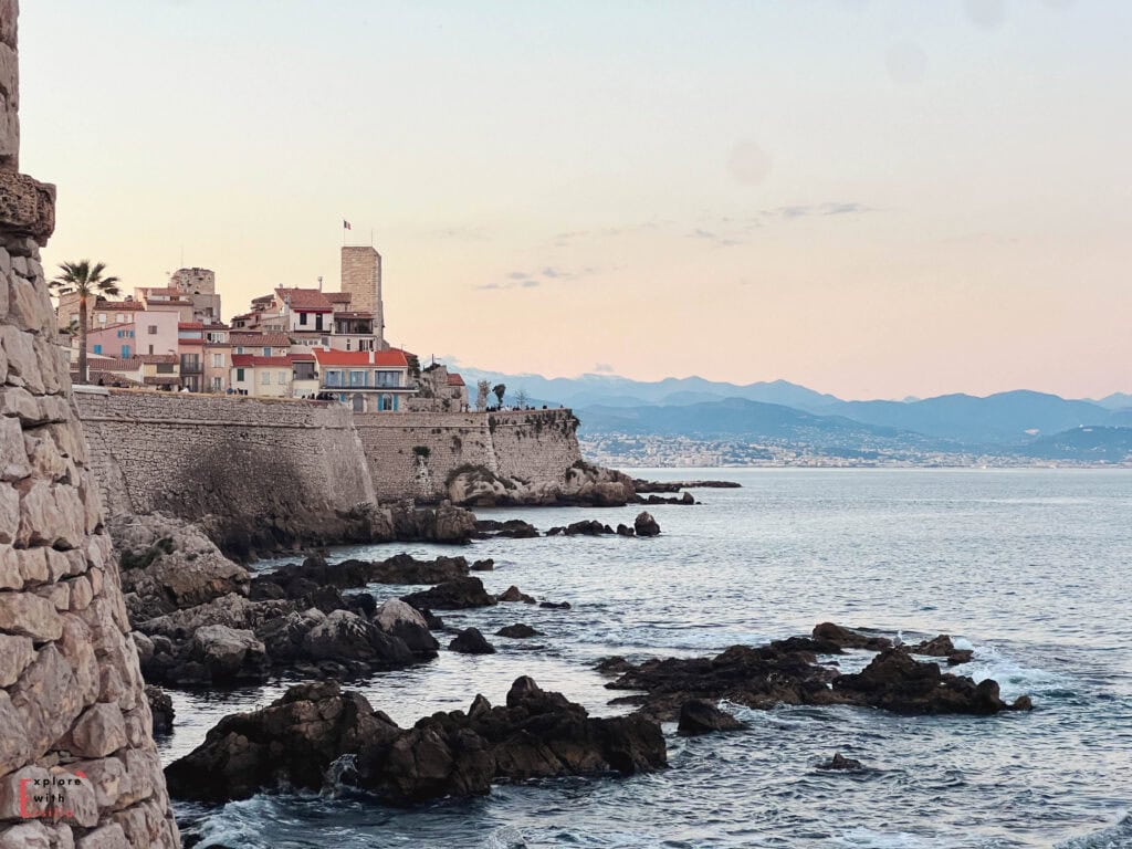 Sunset view of Antibes' Old Town perched on ancient stone ramparts overlooking the Mediterranean Sea, featuring historic buildings with terracotta rooftops, a French flag, rocky coastline with jagged formations in the foreground, and distant mountains silhouetted against a pastel-colored sky.