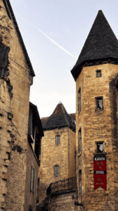 This image captures a historic stone building with pointed slate roofs in Sarlat-la-Canéda, France. A red banner on the right tower reads "Manoir de Gisson - Monument Historique - Entrée," indicating the location is a historic landmark. The medieval architecture, warm-toned stone walls, and intricate window details create a charming old-world atmosphere.