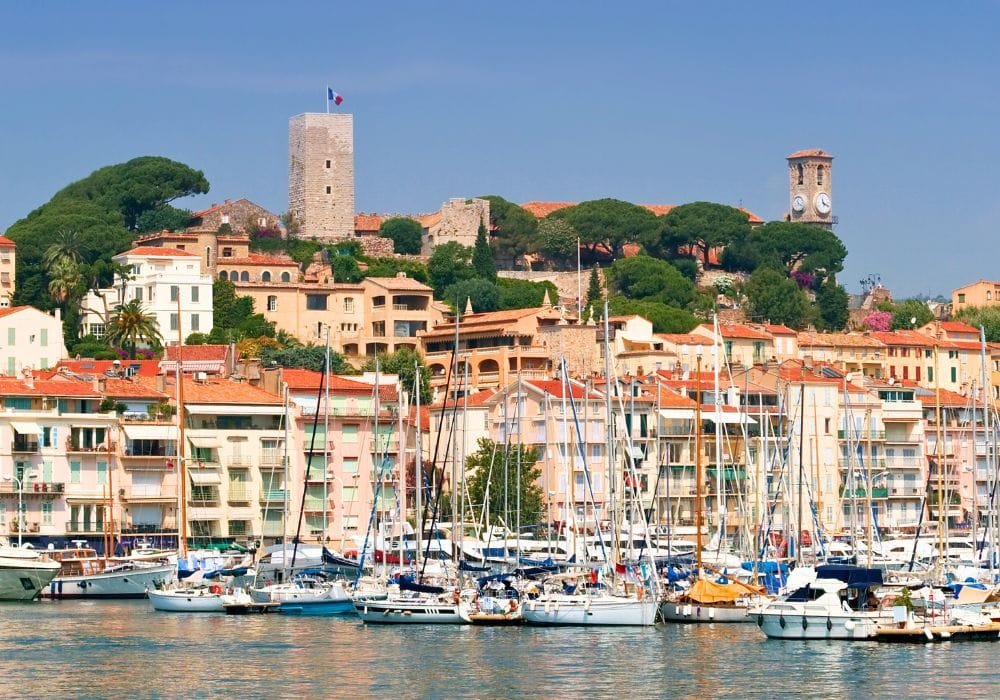 View of Cannes from the harbor showing the historic Le Suquet hillside with its medieval stone watchtower and clock tower rising above pastel-colored buildings with terracotta rooftops, umbrella pine trees, and a marina filled with sailboats and yachts in the foreground, all under bright blue Mediterranean skies.