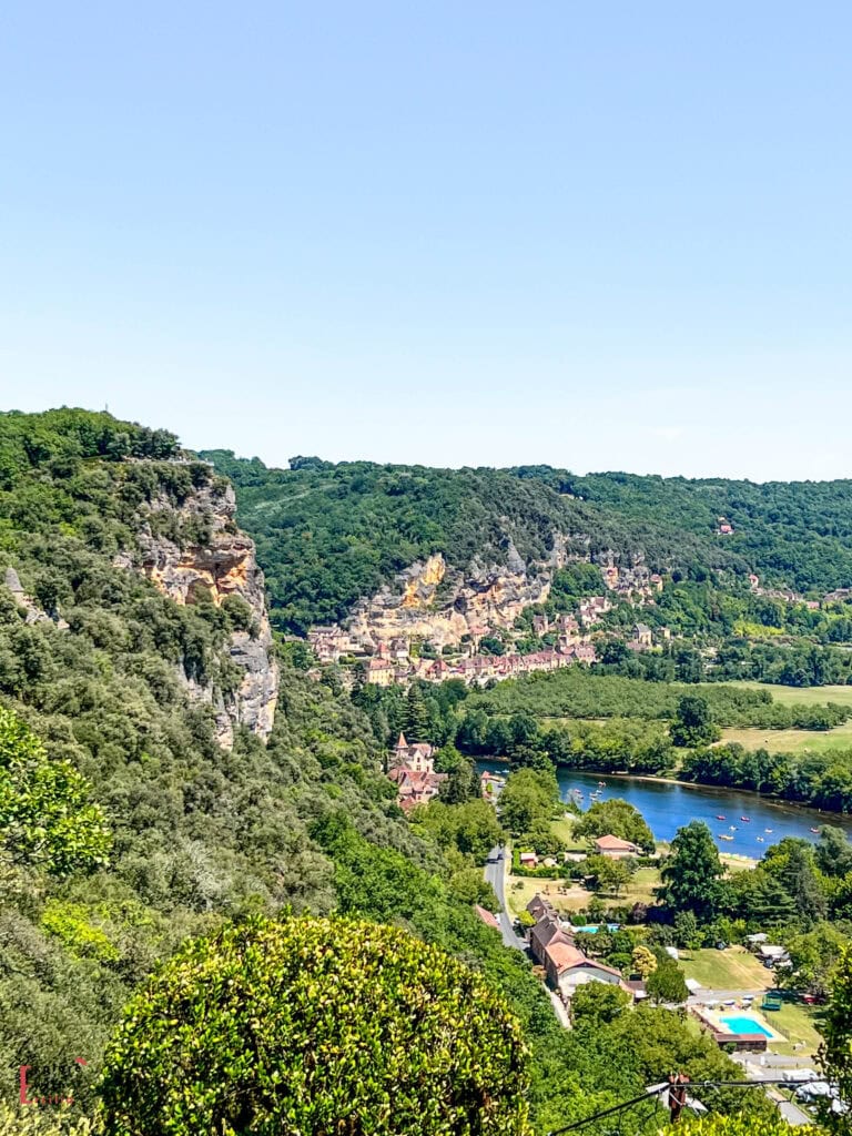 Panoramic view from the Jardins de Marqueyssac lookout point over the Dordogne Valley, showing limestone cliffs, the meandering Dordogne River with kayakers, the village of La Roque-Gageac built into the cliff face, lush green forests, and an outdoor swimming pool visible in the foreground under a clear blue summer sky.