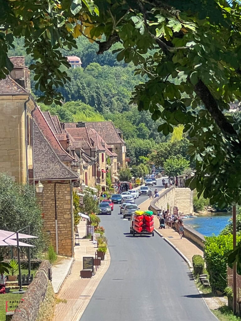 View of La Roque-Gageac village in the Dordogne, showing the main street lined with honey-colored stone buildings with traditional perigordian architecture and terracotta roofs, framed by lush green foliage with the Dordogne River visible alongside the road, and colorful kayaks being transported in the foreground.