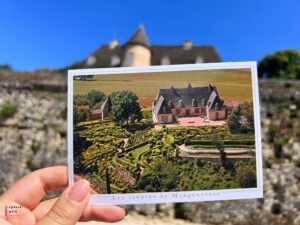 Postcard of Les Jardins de Marqueyssac's ornate topiary gardens and manor house held against backdrop of actual château in Dordogne, France