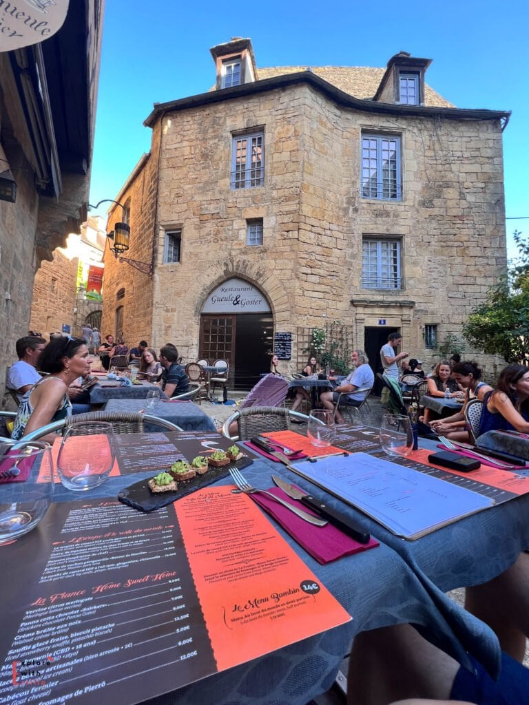 Outdoor dining terrace at Restaurant Gueule & Gosier in Sarlat-la-Canéda, featuring tables set with colorful menus in front of a beautiful honey-colored stone medieval building with a distinctive arched doorway and dormered roof, surrounded by diners enjoying meals under clear blue skies.