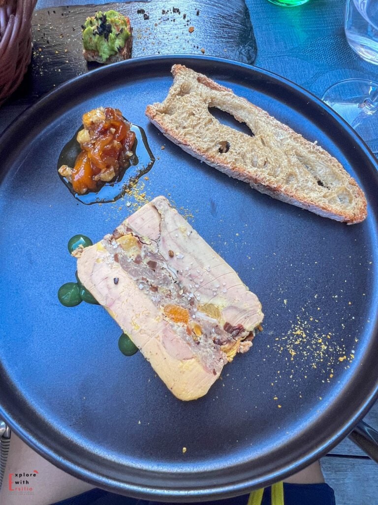 Elegant plate of traditional foie gras terrine served at a restaurant in Sarlat, Dordogne, featuring a slice of duck liver pâté garnished with microgreens, accompanied by toasted artisanal baguette and sweet fruit chutney, presented on a deep blue ceramic plate with avocado toast visible in the background