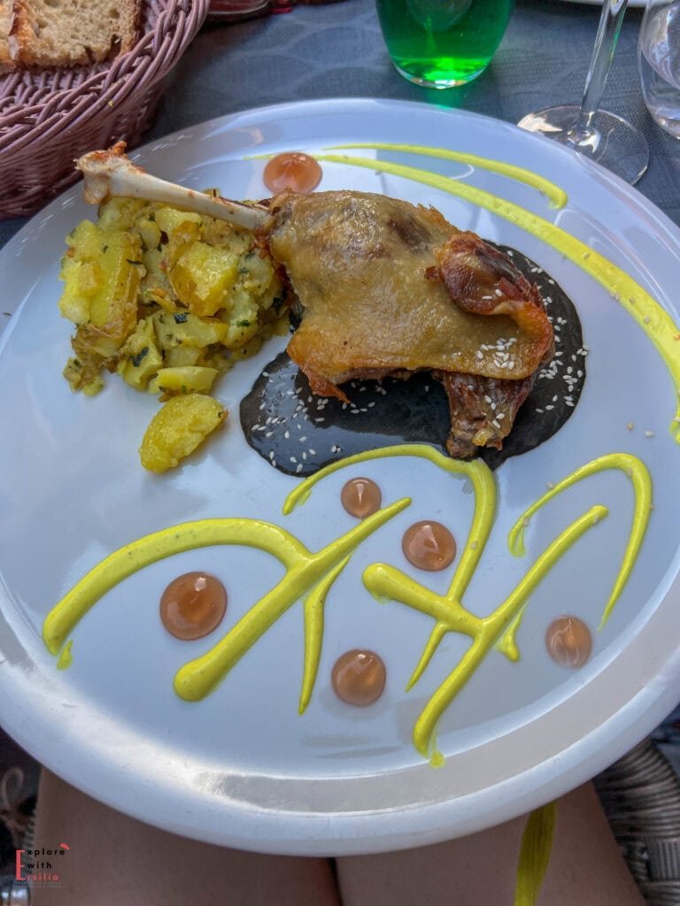 Elegantly presented confit duck leg (confit de canard) served at Restaurant Gueule & Gosier in Sarlat, featuring crispy golden skin atop a black truffle sauce, accompanied by herb-roasted potatoes, and artistically decorated with yellow mustard sauce and amber balsamic pearls on a white plate.