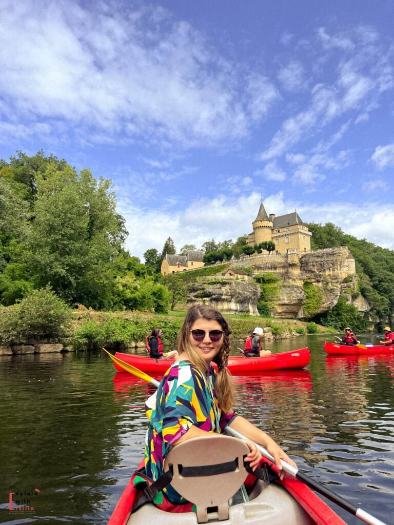 Woman kayaking on the Vézère River in Dordogne with the magnificent Château de Belcayre perched on limestone cliffs in the background, surrounded by lush green forest under bright blue skies with wispy clouds, with other red kayaks visible on the tranquil water.