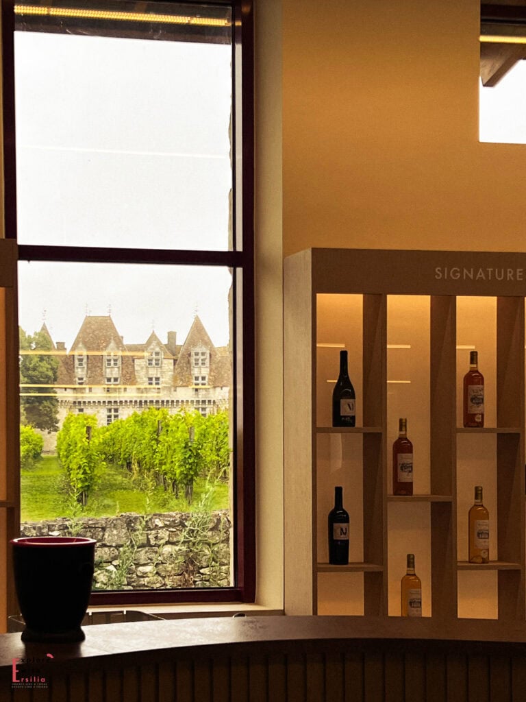 View from inside a wine tasting room at Château de Monbazillac featuring signature wine bottles displayed in illuminated shelving, with a large window framing the Renaissance castle and its surrounding vineyards in the Bergerac wine region of the Dordogne.