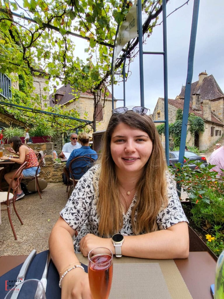 Young woman enjoying a drink at the outdoor terrace of La Petite Tonnelle restaurant in Beynac, with grapevine-covered pergola overhead, surrounded by traditional honey-colored stone buildings with steep roofs, and other diners visible in the charming medieval village setting.