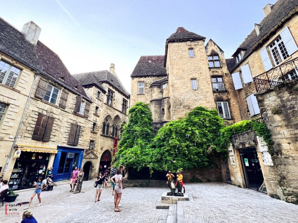 Medieval square in Sarlat's historic center featuring golden limestone buildings with traditional architecture, steep lauze roofs, Gothic windows, and vibrant green ivy, with tourists exploring the cobblestone plaza surrounded by shops and restaurants in this well-preserved Dordogne town