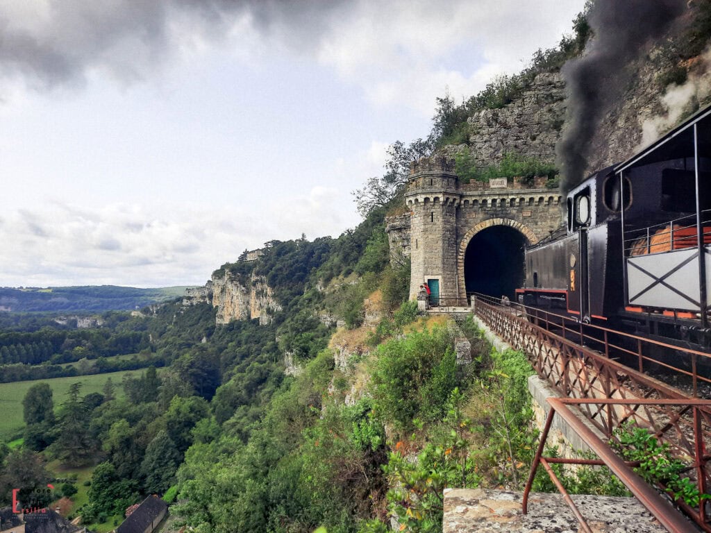 Historic steam train entering a stone tunnel built into a limestone cliff along the scenic Martel Railway near Rocamadour in the Dordogne region, with dramatic views over the lush green valley below, featuring castle-topped cliffs and rolling countryside.