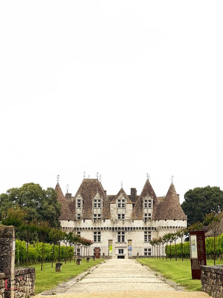 Château de Monbazillac viewed from its vineyard approach, showcasing the distinctive Renaissance architecture with multiple pointed towers, steeply pitched roofs, and cream-colored limestone façade, surrounded by manicured gardens and vineyard rows under an overcast sky in the Dordogne region of France.