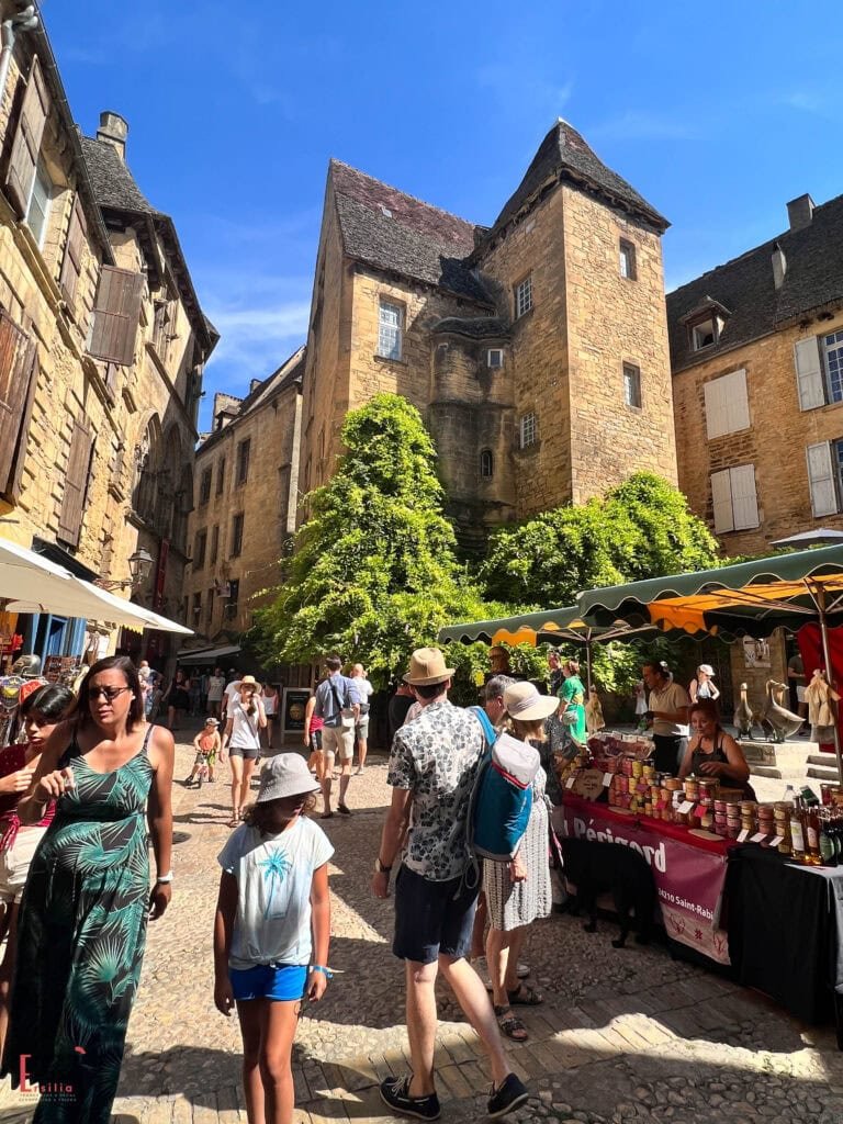 Bustling market day in Sarlat's medieval center with tourists exploring cobblestone streets lined with local product stalls selling regional specialties, surrounded by golden limestone buildings with traditional Perigordian architecture featuring steep roofs and a distinctive tower, all under bright blue summer skies.