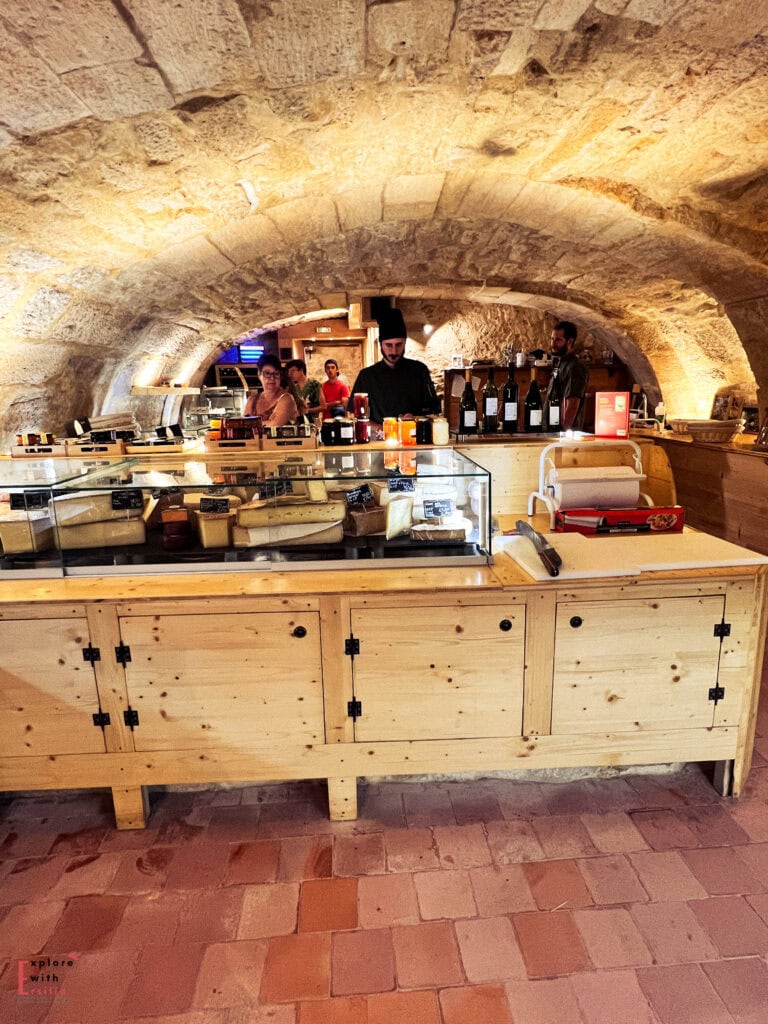 Interior of Chez Pierro cheese shop in Sarlat, Dordogne Valley, France, showing a rustic wooden counter with glass display cases filled with regional cheeses, set inside a traditional stone cave with vaulted limestone ceiling and staff serving customers.