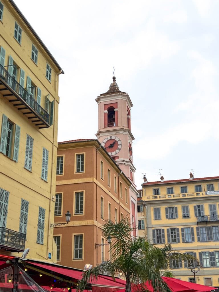 Pink clock tower of the Cathedral of Sainte-Réparate rising above ochre and yellow buildings in Place du Palais de Justice in Nice's Old Town, featuring traditional Provençal shutters, red café awnings, and a palm tree in the foreground, showcasing the Italian-influenced architectural style typical of the historic district.