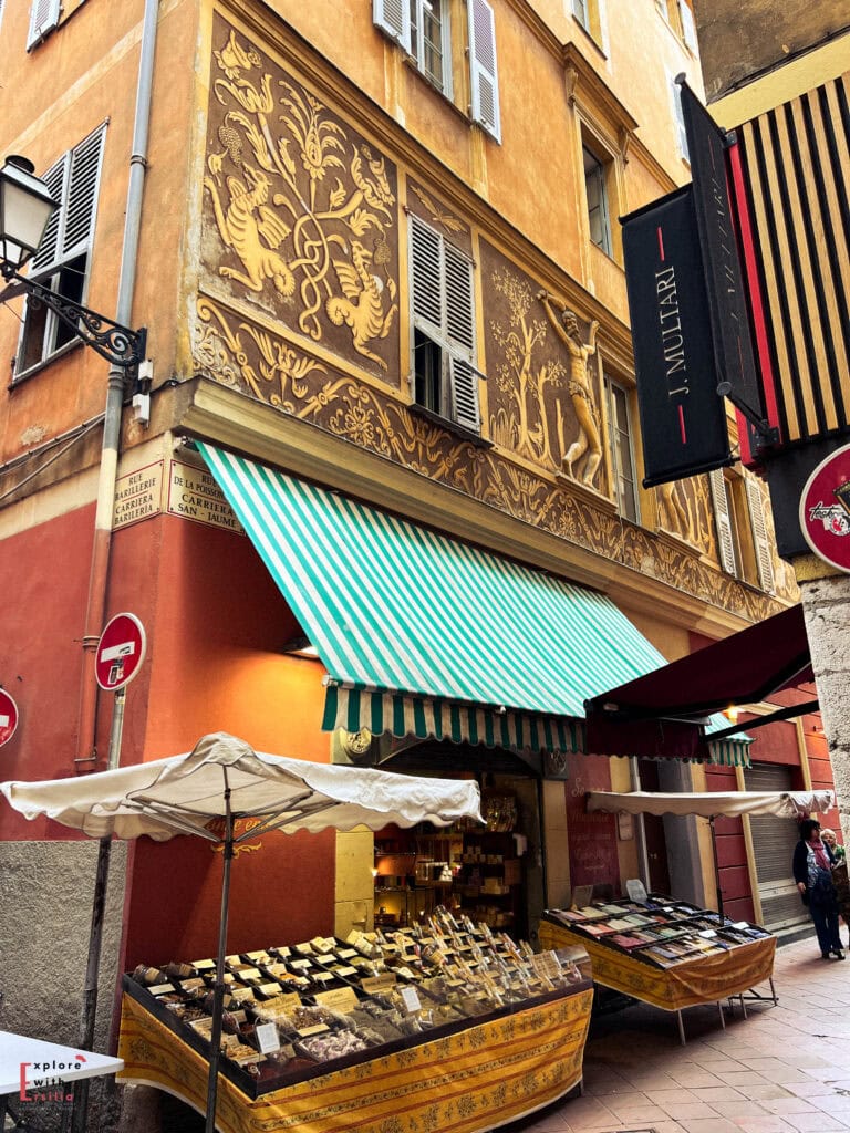 Historic building façade in Nice's Old Town featuring ornate gold sgraffito decorations on an ochre wall, with traditional Provençal shutters, green and white striped awning, and outdoor market stalls displaying local specialty foods beneath, capturing the area's rich Italian architectural heritage.