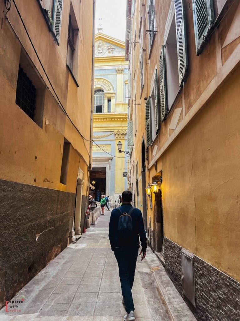 Narrow alleyway in Nice's Old Town with ochre-colored buildings leading toward the yellow Baroque façade of Saint-Jacques-le-Majeur Church, featuring traditional green shutters, warm ambient lighting, and a person walking along the stone-paved street framed by tall historic structures